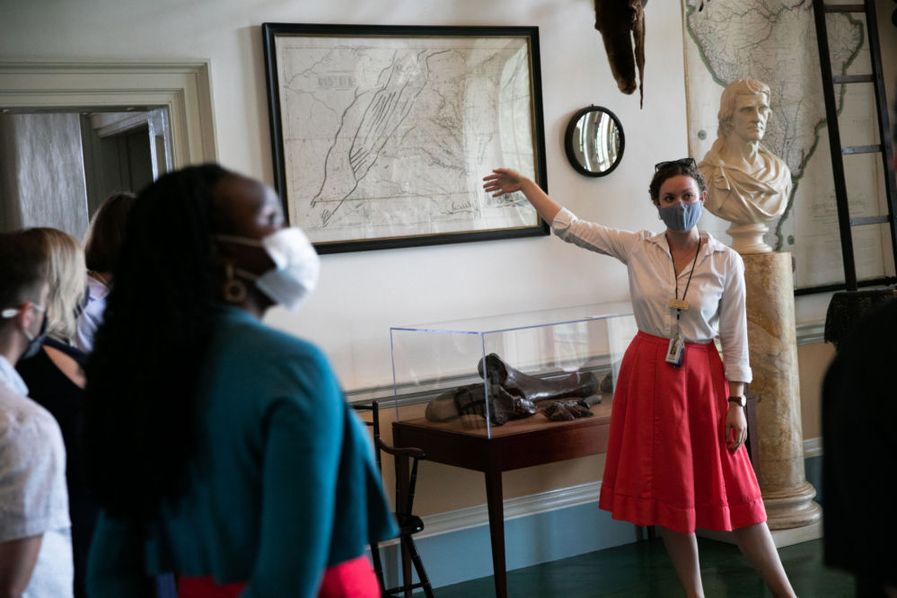 A masked woman points to a map on the wall while others look.