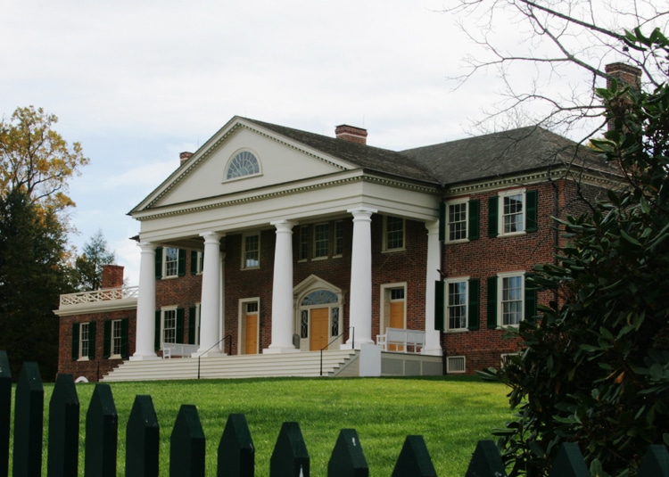 A large brick mansion with white columns behind a green fence.
