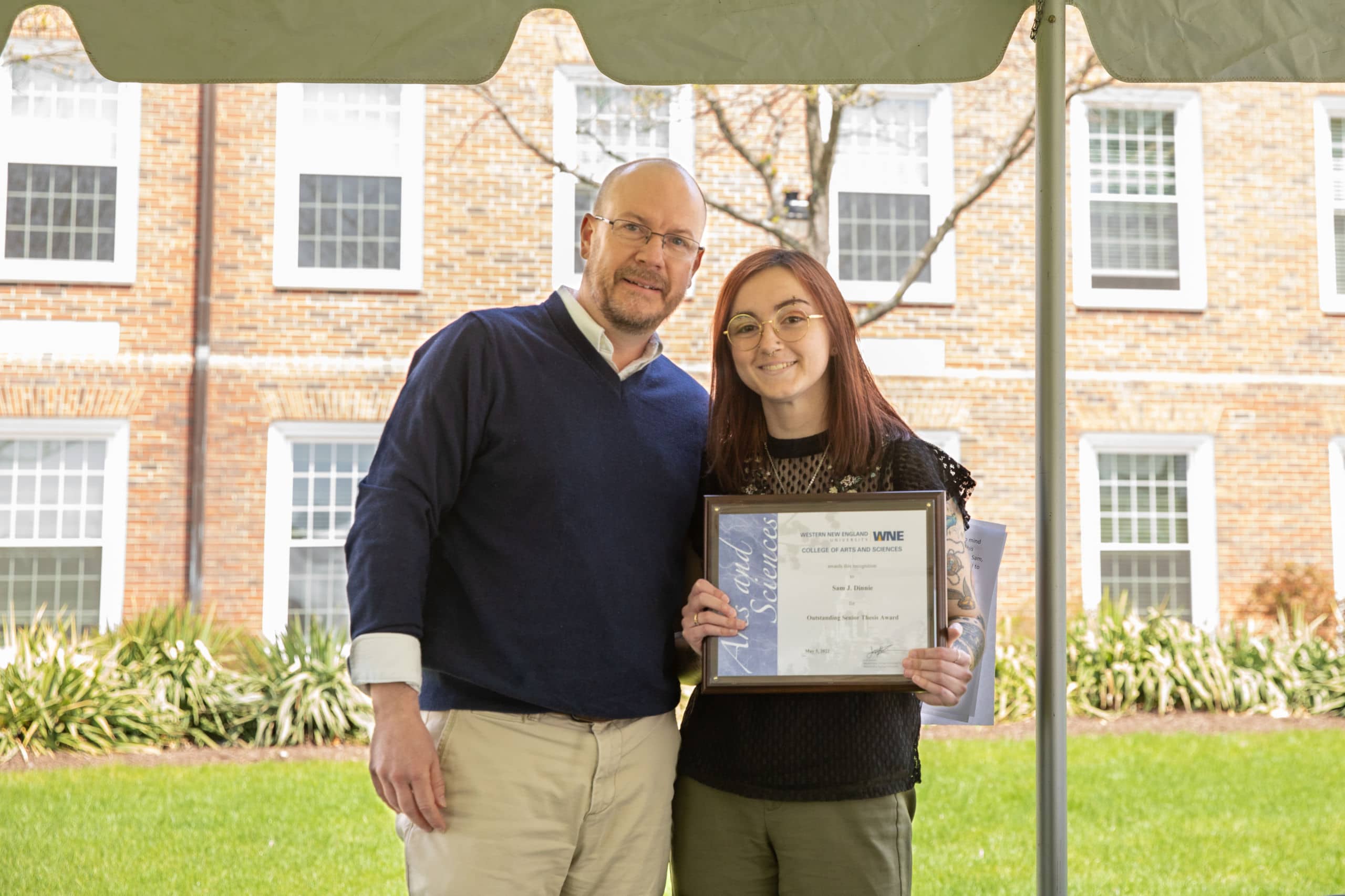Two smiling people holding a certificate outdoors on a college campus.