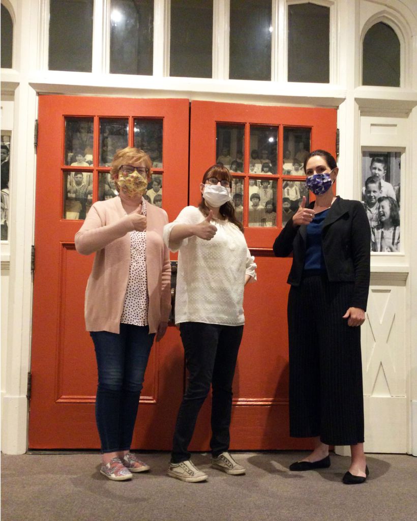 Three masked women giving a thumbs up inside a museum exhibit.