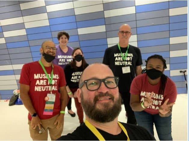 6 smiling people wearing Museums are Not Neutral tshirts taking a selfie.