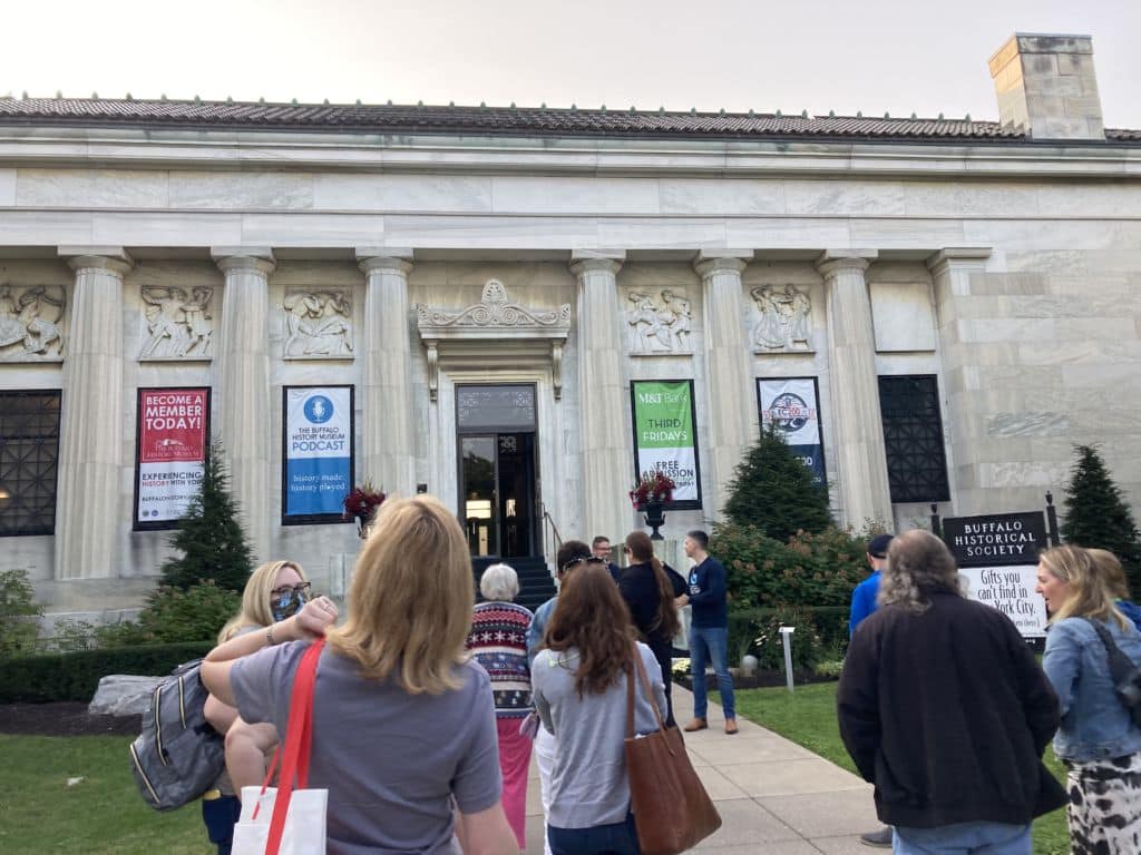 A group of people standing in front of the Buffalo Historical Society, a historic Greek Revival building with columns and Greek scenes.