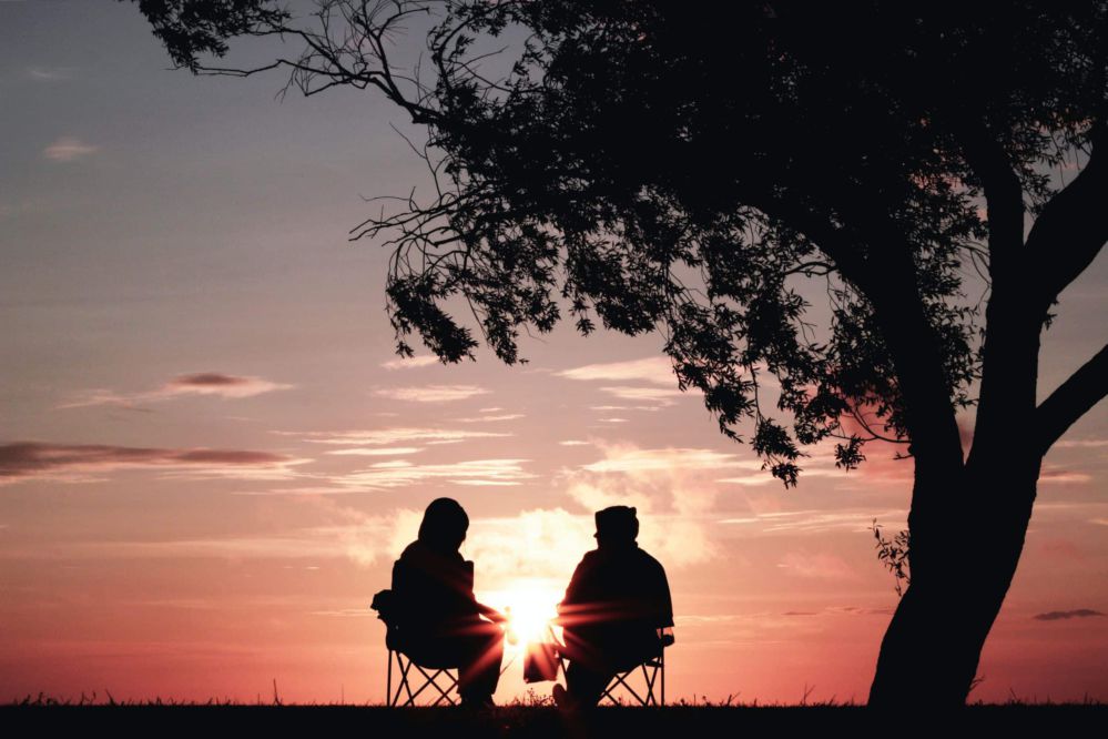 Two silhouetted people sitting on chairs in front of an orange sunset.